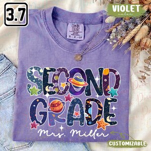 Puede incluir: Una camiseta violeta de manga corta con el texto SECOND GRADE en letras grandes de color azul oscuro rellenas de patrones espaciales que incluyen planetas, estrellas y lunas. Debajo del texto, el nombre Mrs. Miller está impreso en blanco.