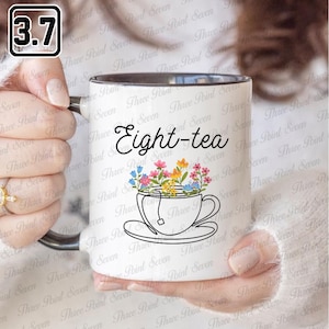 Könnte beinhalten: Weißer Keramikbecher mit schwarzem Innenraum und Henkel, mit dem Text "Eight-tea" und einem floralen Teetassen-Design. Der Becher wird von einer Person gehalten. Ein kleines schwarz-weißes Quadrat mit der Zahl 3,7 befindet sich in der oberen linken Ecke.