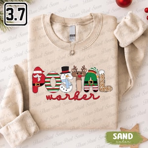 Op de afbeelding: Een zandkleurige sweater met ronde hals en de tekst "Postal worker" in een feestelijk ontwerp. De letters in "Postal" zijn gestileerd als een kerstmanpak, zuurstokstrepen, een sneeuwman, een rendier, een elfenpak en een peperkoekpatroon.