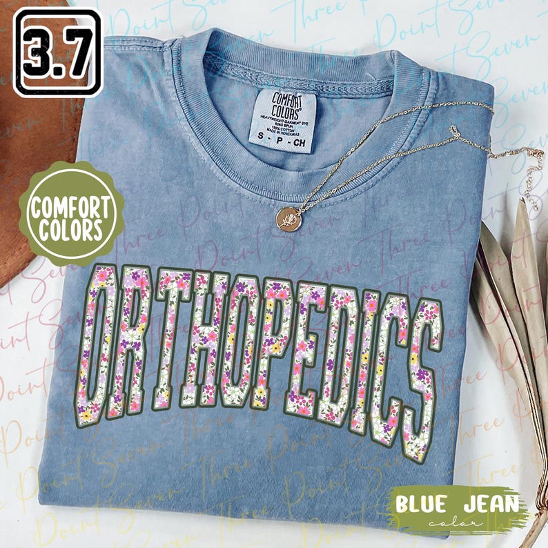 May include: A blue jean t-shirt with the word "ORTHOPEDICS" in white letters outlined in green. The letters are covered in a floral pattern with pink, purple, and yellow flowers. The shirt has a tag that says "COMFORT COLORS".