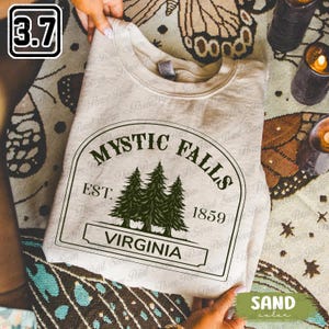 May include: A sand-coloured crew neck sweatshirt. The front displays a black and white graphic with the words "MYSTIC FALLS" and "VIRGINIA" and the year "1859". The sweatshirt is folded and presented on a patterned surface.