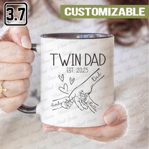 Può includere: Tazza in ceramica bianca con interno e manico neri. La tazza presenta la scritta "TWIN DAD EST. 2025" con una grafica di mani che si tengono. I nomi "Lindy" e "Bella" sono scritti sulle mani. La parola "Dad" è scritta anche sulla mano.