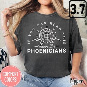May include: A dark grey t-shirt with a white graphic design that reads "If you can read this - Thank the Phoenicians". The graphic includes a stylised globe with mountains and plus signs.