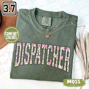 May include: A green t-shirt with a floral print of the word "DISPATCHER" in pink, purple, and yellow. The shirt is made by Comfort Colors and has a moss color.
