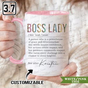 Puede incluir: Taza de cerámica blanca de 425 ml con interior y asa rosas. La taza presenta el texto "BOSS LADY" en una mezcla de fuentes y colores, junto con una definición. La taza es personalizable.
