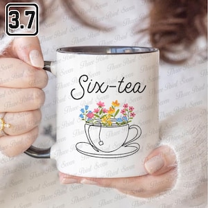 May include: White ceramic mug with a black interior and handle. The mug features the text "Six-tea" in black script above a floral teacup design. The teacup is filled with colorful flowers.