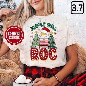 Może przedstawiać: Kremowa koszulka z grafiką świąteczną. Projekt zawiera napis "JINGLE BELL ROC" z czapką Świętego Mikołaja, choinkami i płatkami śniegu. Koszulka posiada również logo Comfort Colors.