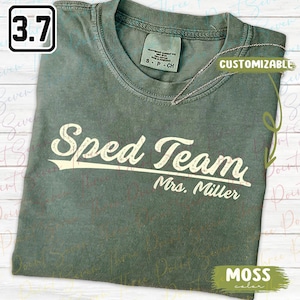May include: A moss green crew neck t-shirt featuring the white text Sped Team and Mrs. Miller in a vintage-style font. The shirt has a distressed graphic design, a ribbed collar, and a neck label indicating size S-CH, made of 100% cotton.