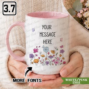 May include: A white ceramic mug with a pink interior and handle, featuring a colourful floral pattern at the base and the black text "YOUR MESSAGE HERE" in the centre. The mug includes small butterfly illustrations and has a 15 oz capacity.