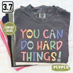 May include: A folded dark grey t-shirt featuring the phrase "YOU CAN DO HARD THINGS!" in colourful, retro lettering. The shirt is part of a stack of other coloured t-shirts. The label reads "Comfort Colors".