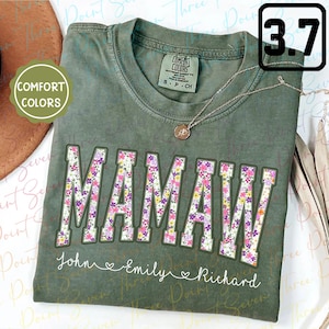 May include: A green t-shirt with the word "MAMAW" in large white letters outlined in pink. The letters are filled with a floral pattern. The text "John Emily Richard" is written below the word "MAMAW" in a smaller font.