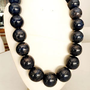 May include: A dark blue beaded necklace displayed on a white bust. The beads are large, round, and have a glossy finish with subtle variations in color. The necklace is a statement piece, likely for fashion or jewelry.