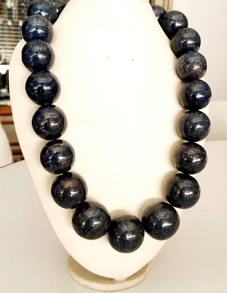 May include: A dark blue beaded necklace displayed on a white bust. The beads are large, round, and have a glossy finish with subtle variations in color. The necklace is a statement piece, likely for fashion or jewelry.