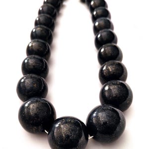 May include: A necklace featuring large, round, dark beads with a metallic sheen. The beads are strung together with a silver-colored clasp. The necklace is displayed against a white background.