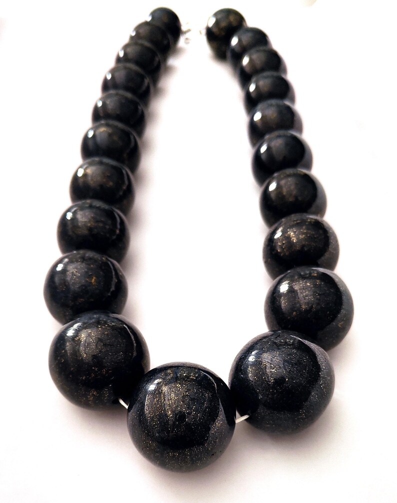 May include: A necklace featuring large, round, dark beads with a metallic sheen. The beads are strung together with a silver-colored clasp. The necklace is displayed against a white background.