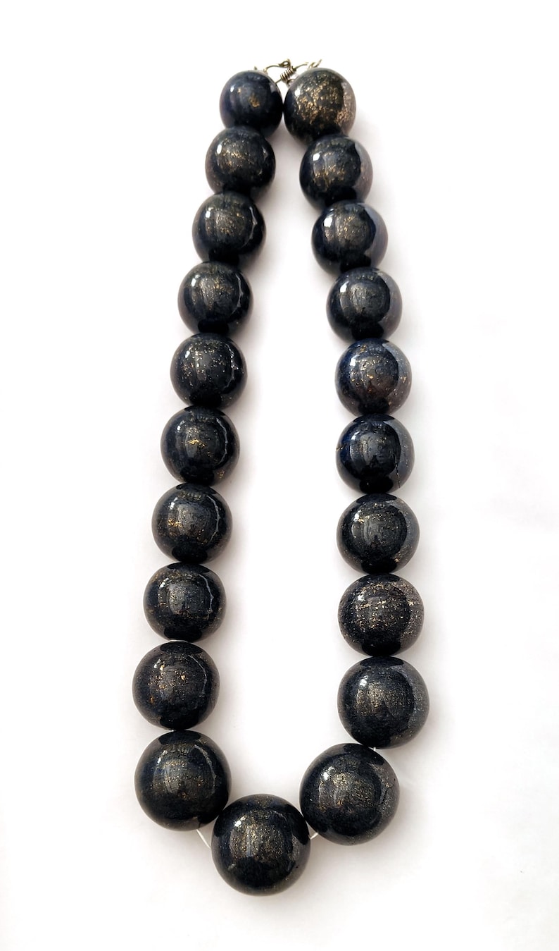 May include: A necklace featuring large, round beads in shades of dark blue and black with a speckled, metallic gold pattern. The beads are strung together with a silver clasp, creating a striking jewelry piece.