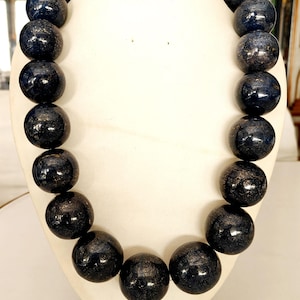 May include: A dark blue beaded necklace displayed on a white bust. The beads are large, round, and have a glossy finish with subtle variations in color. The necklace is a statement piece, likely for fashion or special occasions.