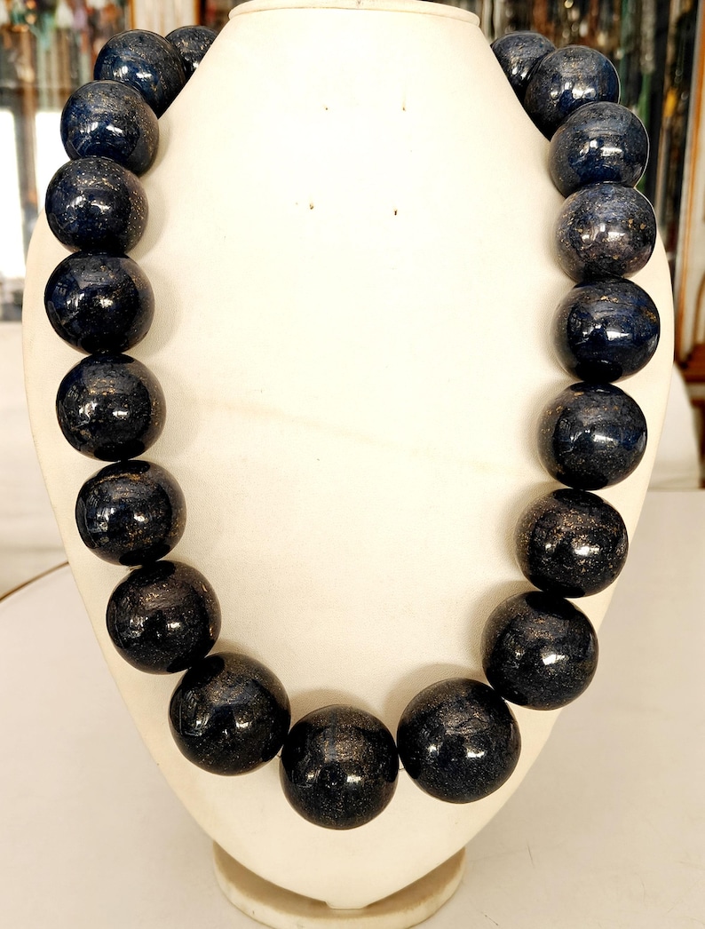 May include: A dark blue beaded necklace displayed on a white bust. The beads are large, round, and have a glossy finish with subtle variations in color. The necklace is a statement piece, likely for fashion or special occasions.