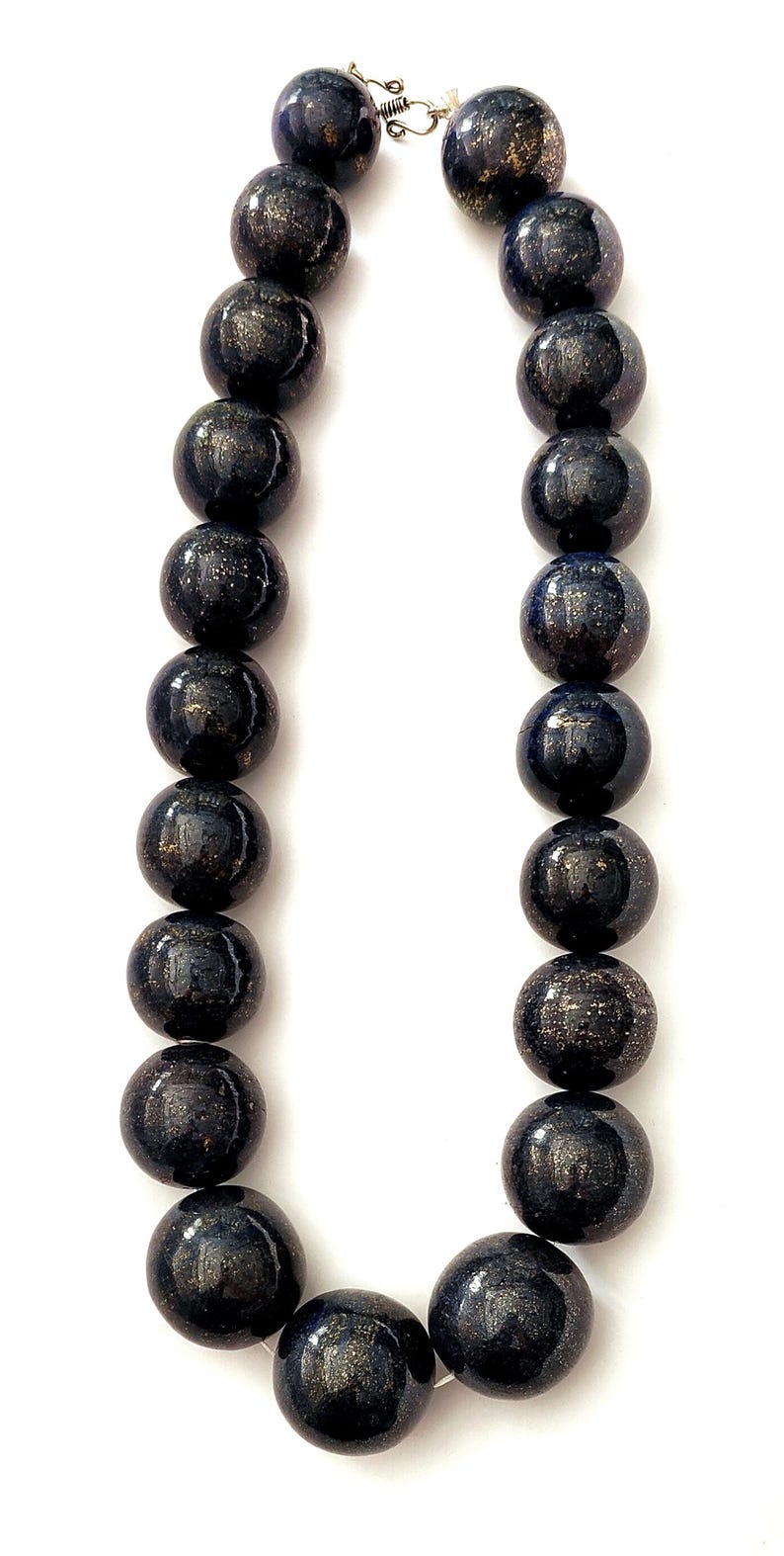 May include: A necklace featuring large, round, dark blue beads with gold flecks. The beads are strung together with a silver clasp. The necklace is displayed against a white background.