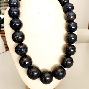 May include: A dark blue beaded necklace with a metallic sheen, displayed on a white jewelry bust. The necklace features large, round beads, creating a bold and eye-catching accessory. The beads have a glossy finish.