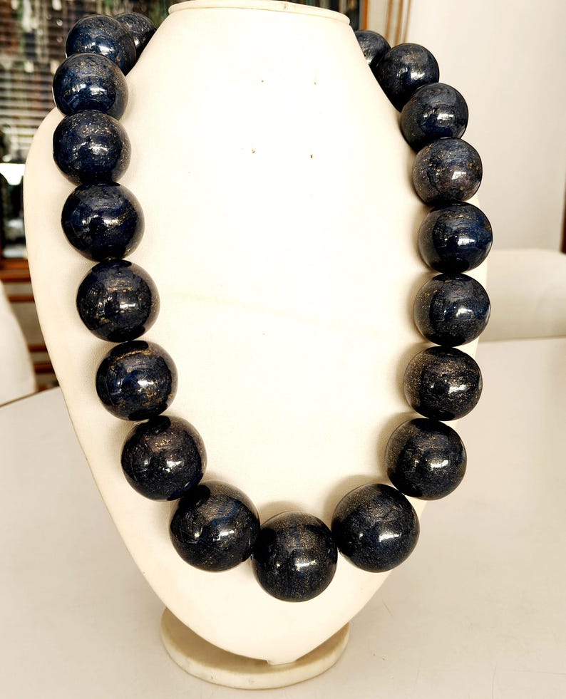 May include: A dark blue beaded necklace with a metallic sheen, displayed on a white jewelry bust. The necklace features large, round beads, creating a bold and eye-catching accessory. The beads have a glossy finish.