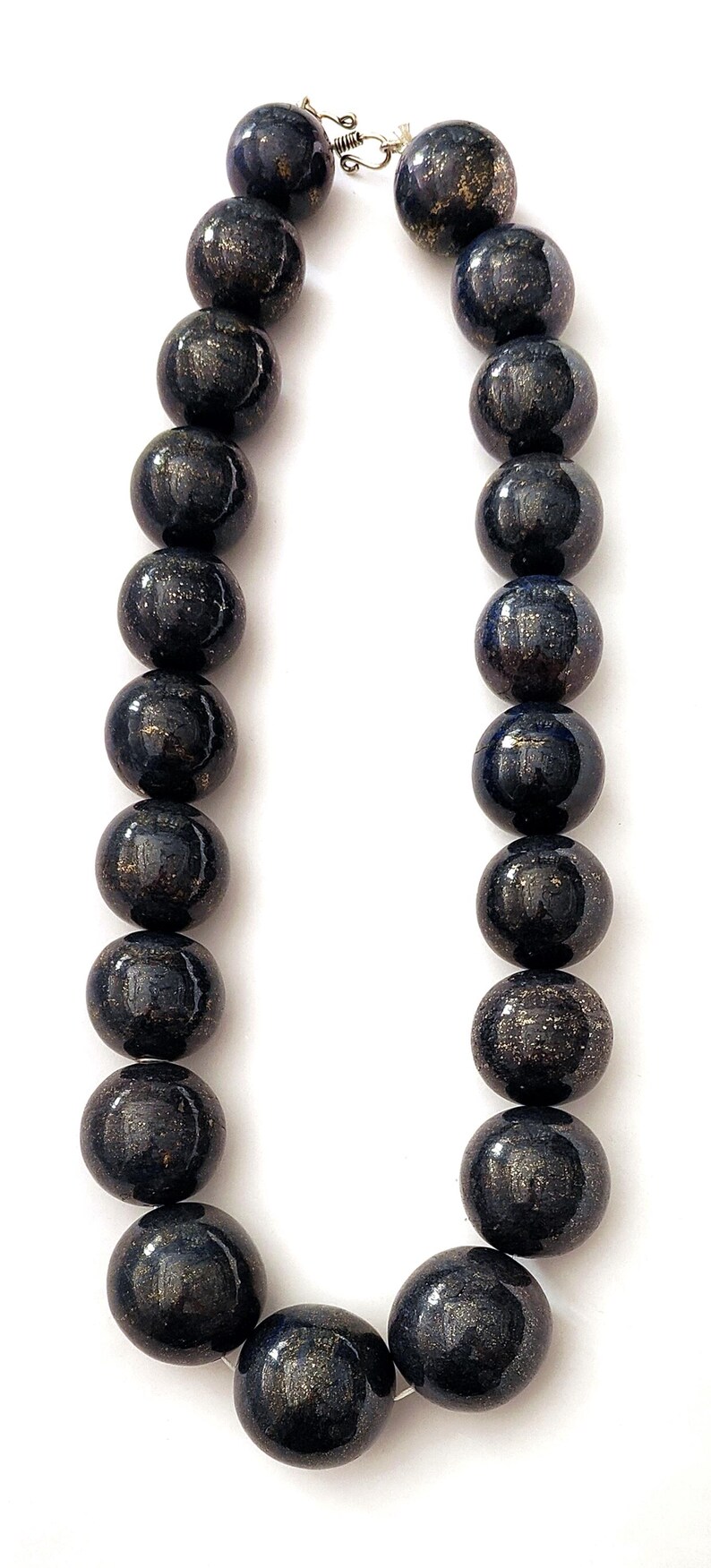 May include: A necklace featuring large, round beads in a dark blue color with gold flecks. The beads are strung together with a silver clasp. The necklace is displayed against a white background.