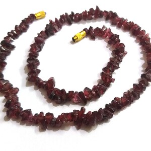 May include: A necklace made of small, irregular, dark red garnet chips. The necklace is strung on a thin cord and has gold-coloured clasps.