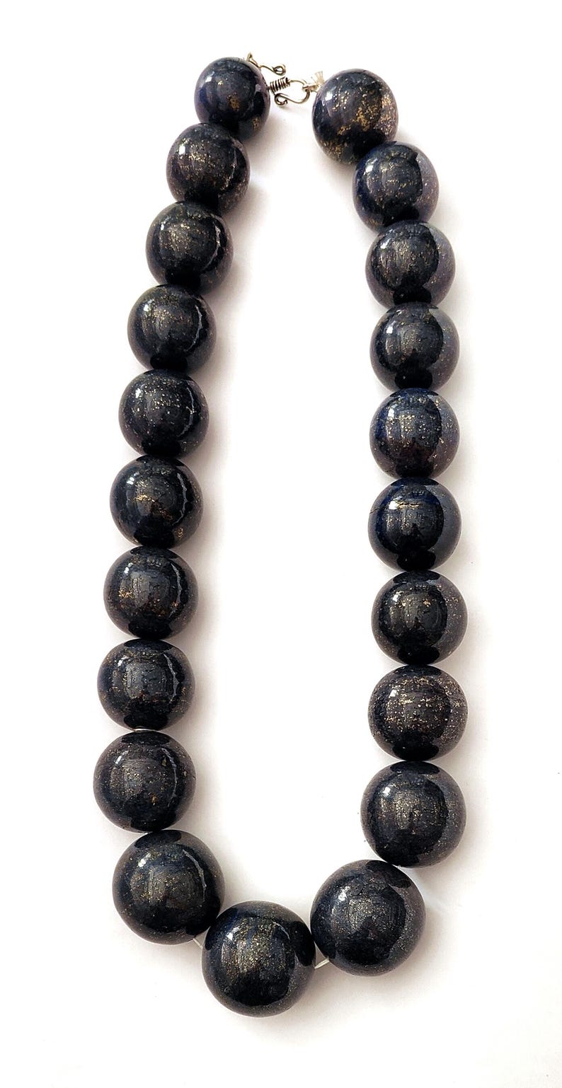 May include: A necklace featuring large, round beads in a dark navy blue color with gold flecks. The beads are strung together with a silver clasp. The necklace is displayed against a white background.
