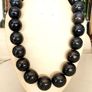 May include: A dark blue beaded necklace displayed on a white bust. The beads are large, round, and have a speckled, metallic appearance. The necklace is a statement piece, likely for fashion or special occasions.