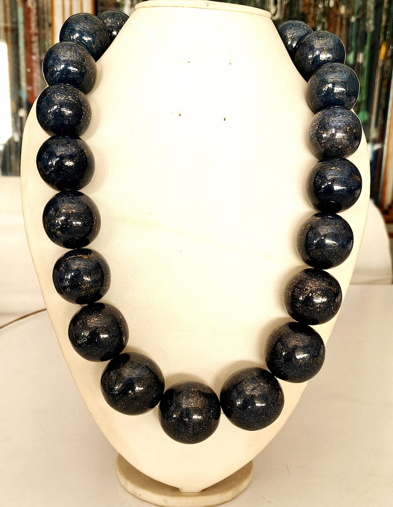 May include: A dark blue beaded necklace displayed on a white bust. The beads are large, round, and have a speckled, metallic appearance. The necklace is a statement piece, likely for fashion or special occasions.