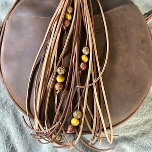 May include: A brown leather round crossbody bag with a long leather tassel. The tassel features strands of light and dark brown leather, adorned with yellow, brown, and gold beads. The bag has a strap and a top closure.