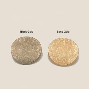 May include: Two circular metal buttons with a textured, brushed finish. The button on the left is labeled Black Gold and features a dark, metallic tone. The button on the right is labeled Sand Gold and has a lighter, warm metallic appearance.