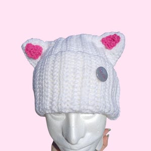 May include: A white crocheted beanie with pink inner ears and a small silver button that says "Mango". The hat is displayed on a white mannequin head against a pink background. The hat is held by a hand.