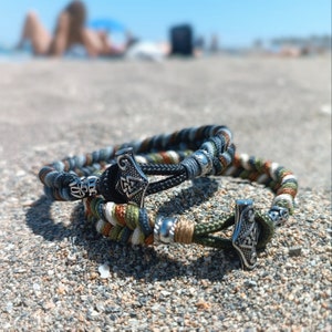 Può includere: Due braccialetti intrecciati con dettagli in metallo argentato. I braccialetti sono realizzati con corda verde, marrone e bianca. I dettagli in metallo hanno la forma di un martello.