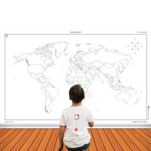 May include: A black and white world map poster with a compass in the top right corner. The poster is hanging on a wall with wood paneling. A child is standing in front of the poster, looking at it.