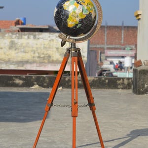 Puede incluir: Un globo terráqueo de estilo vintage con un diseño de océano negro y continentes coloridos. Está montado sobre un trípode de madera. El texto "Nautical Collection" es visible. El trípode es de madera.