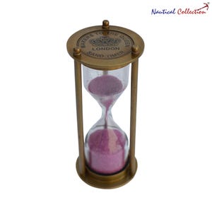 May include: A brass hourglass with a clear glass chamber containing pink sand. The top is engraved with "MAKERS TO THE QUEEN LONDON SAND TIMER" and a crown. The hourglass is a decorative item, measuring approximately 15 cm in height.
