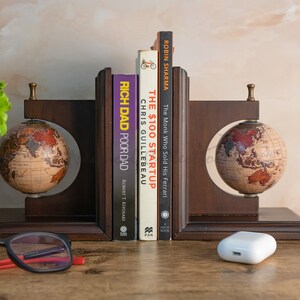 Op de afbeelding: Een paar houten boekenleggers met globes aan elke kant. De boekenleggers ondersteunen een verzameling boeken, waaronder "Rich Dad Poor Dad" van Robert Kiyosaki, "The $100 Startup" van Chris Guillebeau en "The Monk Who Sold His Ferrari" van Robin Sharma.
