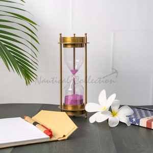 May include: A brass hourglass with a purple sand timer, a notebook with a pen, and two white plumeria flowers on a dark wood surface. The background is a white tiled wall.