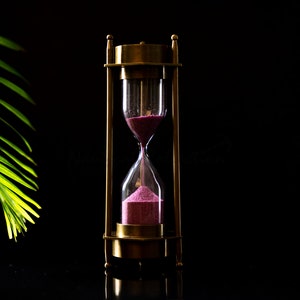 May include: A brass hourglass with pink sand, set against a black background. The sand is mostly in the bottom bulb, with a small amount in the top bulb.