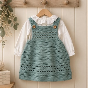 Puede incluir: Un vestido tipo pichi de ganchillo en color verde salvia con un patrón de punto texturizado, bandas horizontales caladas y dos botones de madera en los tirantes, sobre una blusa blanca de manga larga con un delicado cuello de encaje.