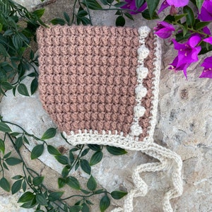 Op de afbeelding: Een gehaakte baby muts in een lichtbruine kleur met een witte gehaakte rand en witte pompoenen. De muts heeft een lange witte gehaakte strik.