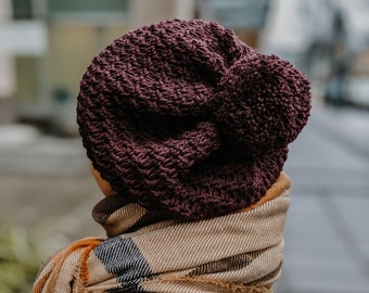 May include: A dark purple knitted beanie with a fluffy pom pom on top. The beanie is worn by a person wearing a plaid scarf.
