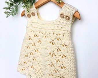 Pode incluir: Um vestido branco de crochê com uma bainha festoneada e detalhes de botões. O vestido está pendurado em um cabide de madeira.