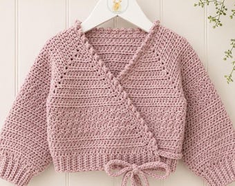 May include: A dusty rose colored long-sleeve wrap cardigan made of crocheted yarn. The garment features a V-neckline, ribbed cuffs, and a matching crocheted tie closure at the waist. The cardigan is displayed on a white plastic hanger.
