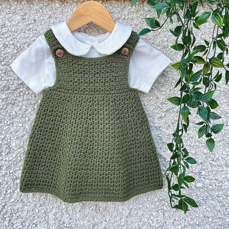 May include: A green crocheted dress with a white Peter Pan collar and two small buttons. The dress is on a white textured background.