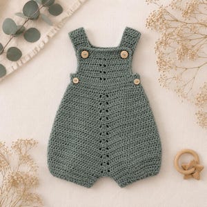 May include: A sage green crochet romper featuring a central vertical decorative stitch pattern, wide shoulder straps, and four light-colored wooden buttons. The garment has a rounded neckline and leg openings, showcasing a textured yarn construction.