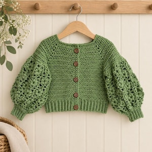May include: A sage green crochet cardigan featuring long, voluminous sleeves with a detailed floral pattern. The garment has a ribbed hem and cuffs, a round neckline, and a front button closure with five circular brown wooden buttons.
