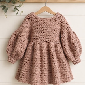 Könnte beinhalten: Ein altrosa gehäkeltes Kleid mit rundem Ausschnitt, einem eng anliegenden Oberteil und voluminösen Puffärmeln mit gerippten Bündchen. Der Rock des Kleides ist an der Taille gerafft, was eine ausgestellte Silhouette mit einem strukturierten Muster erzeugt.