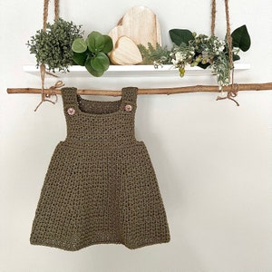 May include: A green crocheted dress with two buttons on the straps. The dress is hanging on a wooden branch with greenery on either side.